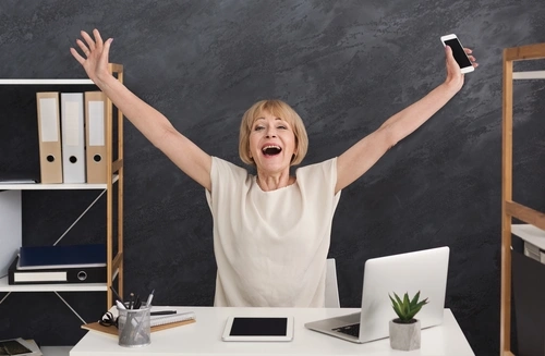 женщина с гипоманиией сидит за столом подняла руки и радуется